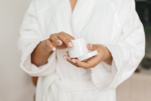 Woman in a bathrobe holding a skincare cream jar in an indoor setting.